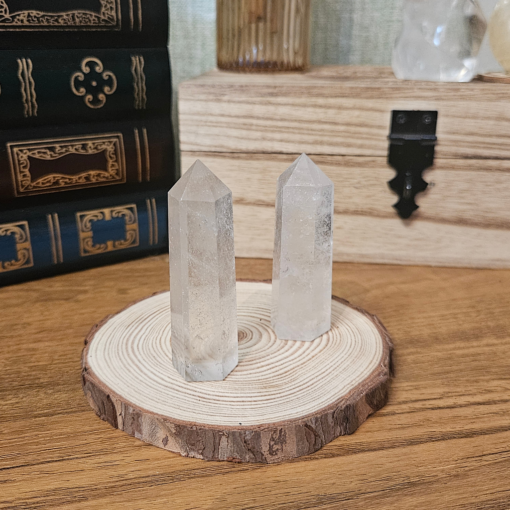 Two clear crystal point towers displayed on a round natural wood slice coaster, set on a wooden tabletop with books and a wooden chest in the background.