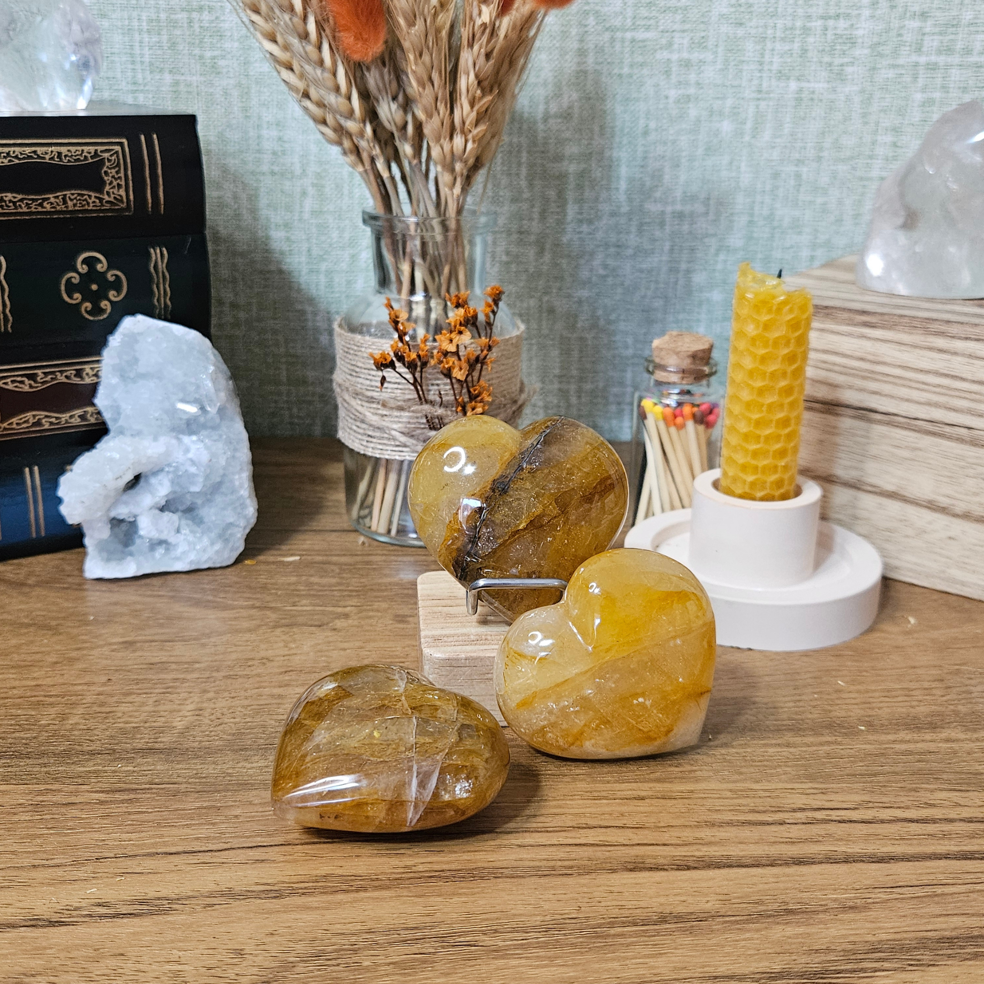 Three 50mm Golden Healer Quartz hearts with natural honey, amber and golden inclusions displayed with dried flowers and a beeswax candle.